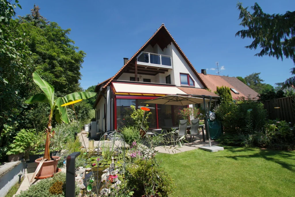 Wintergarten in Ahorntal unter dem Balkon eines Hauses mit Satteldach, weinrote Profile und Markise, Außenansicht
