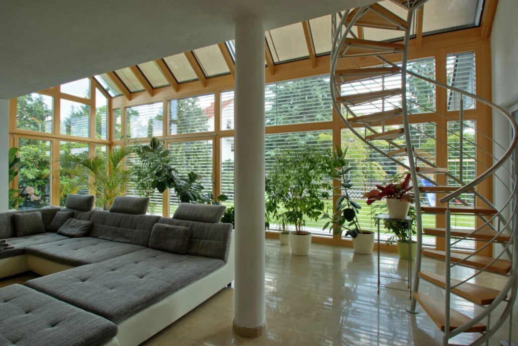 Glasanbau in Bruck innen mit grauer Wohnlandschaft, weißer Stütze, Wendeltreppe, Pflanzen und Holz-Glasfront mit Raffstores
