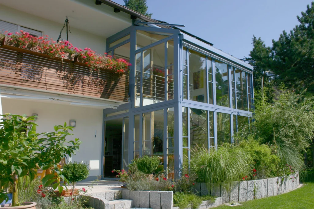 Zweigeschossiger Wintergarten in Bubenreuth mit blaugrauen Rahmen und Glasdach um den Balkon eines Hauses