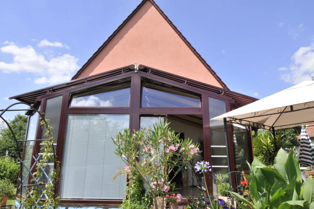 Wintergarten in Burk mit fünfeckigem Vorbau, Glasdach und dunkelroter Aluminiumschale, Blick von der Terrasse