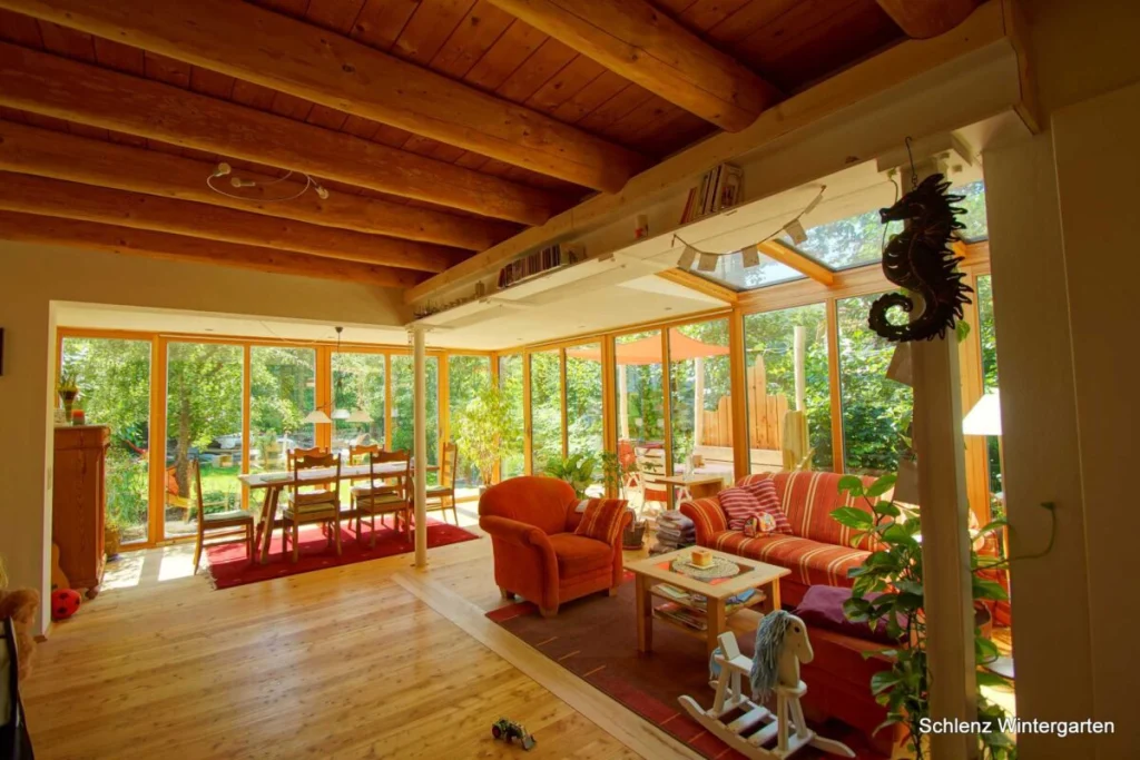 Anbau in Grafenau innen mit Holzbalkendecke, roten Sesseln, Esstisch und raumhoher Verglasung zum Garten