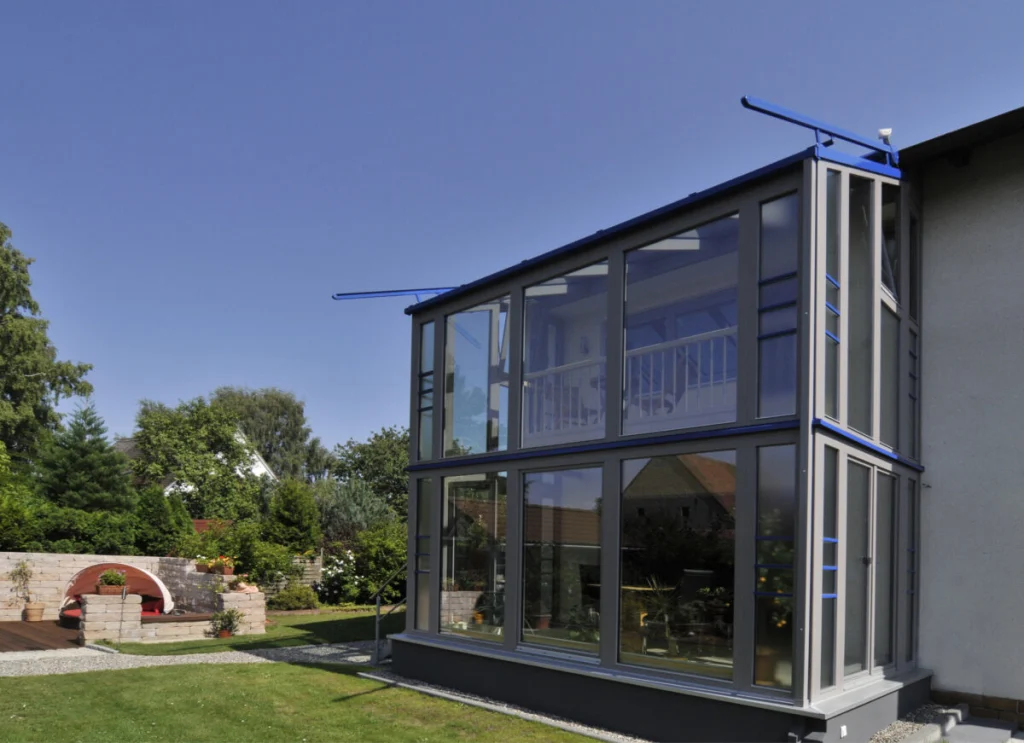 Wintergarten in Hannberg von außen, zweigeschossiger Glaskubus mit grauen Profilen und blauen Details am Wohnhaus