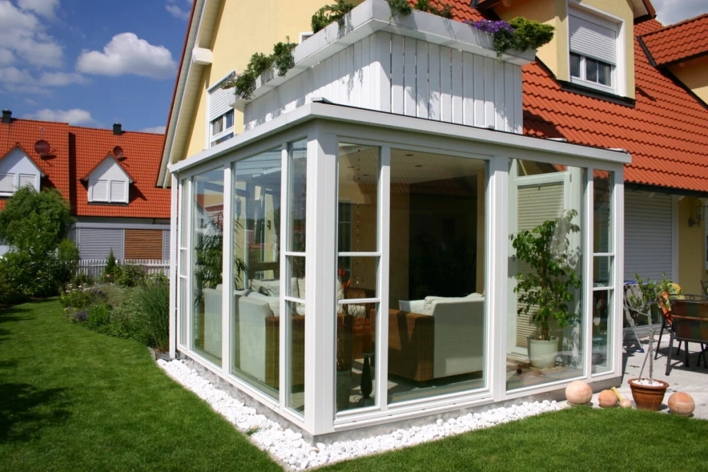 Wintergarten in Hausen unter dem Eckbalkon eines gelben Hauses, weiße Profile, Kiesstreifen und Rasen