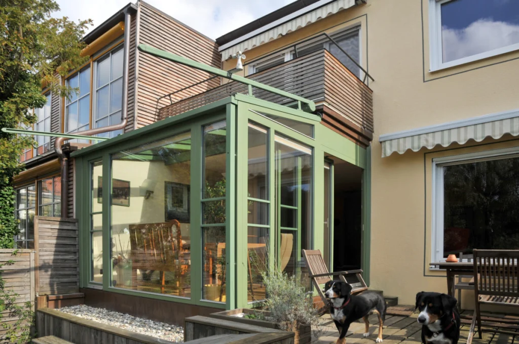 Wintergarten in Schoppershof mit grünen Rahmen und Glasdach unter dem Balkon, davor zwei Hunde auf der Terrasse