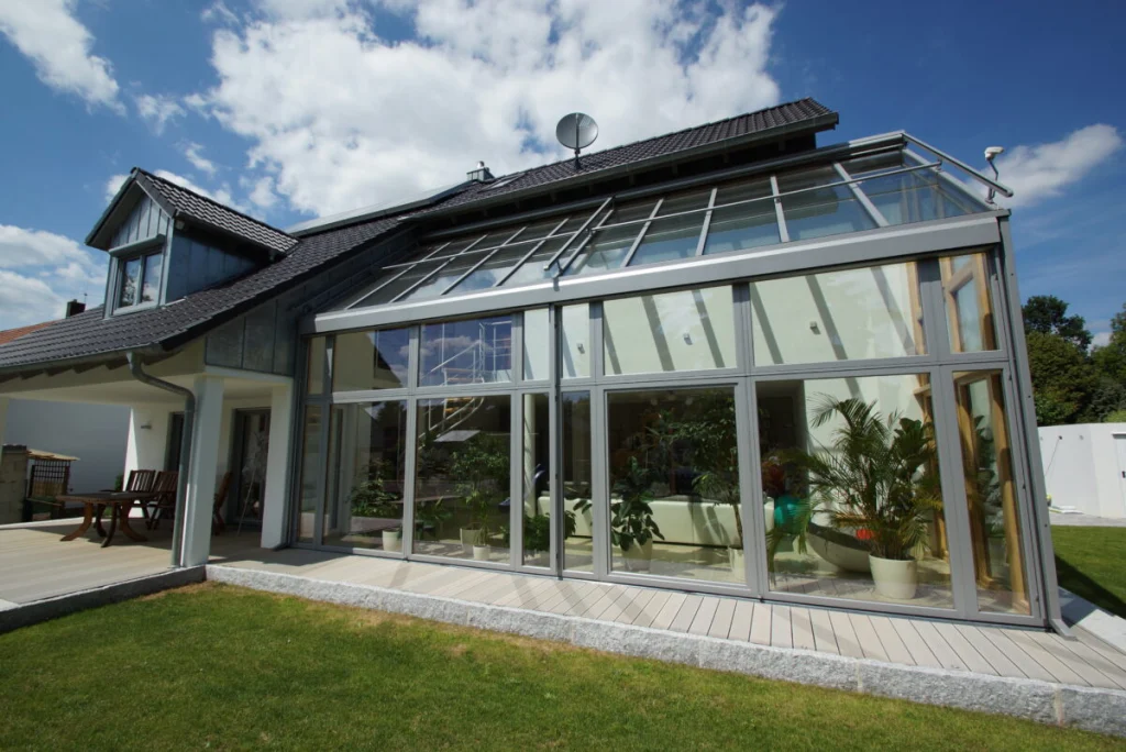 Glasanbau in Bruck von der Seite mit Glasdach, hellgrauen Rahmen, überdachter Terrasse und Holzdeck