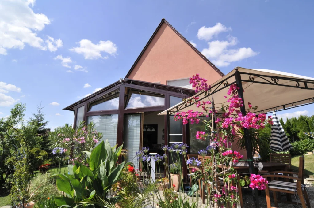 Wintergarten in Burk mit Glasdach und dunkelroten Rahmen zwischen Bougainvillea und Gartenpavillon im Sommer
