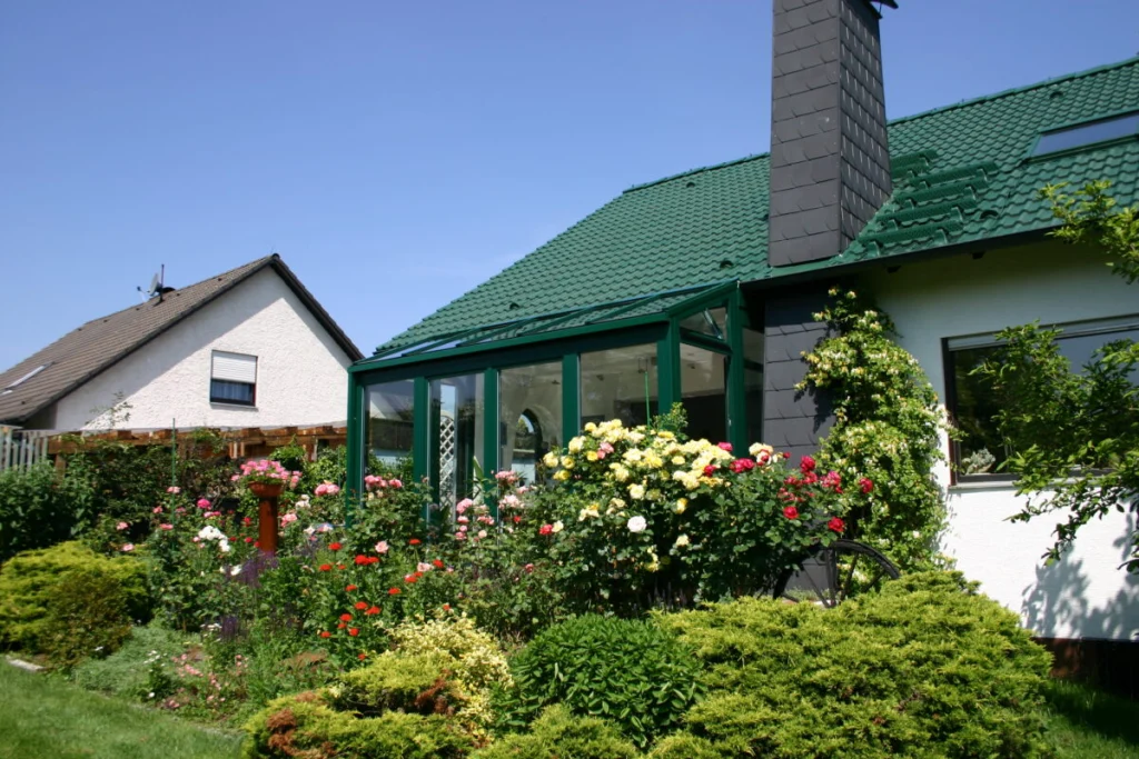 Wintergarten in Effeltrich mit dunkelgrünen Rahmen und Glasdach an einem Haus mit grünem Ziegeldach und Rosengarten