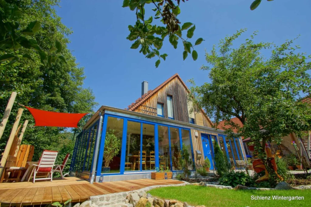 Flachdachanbau in Grafenau vom Garten mit blauen Rahmen, rotem Sonnensegel und Holzdeck mit Liegestühlen