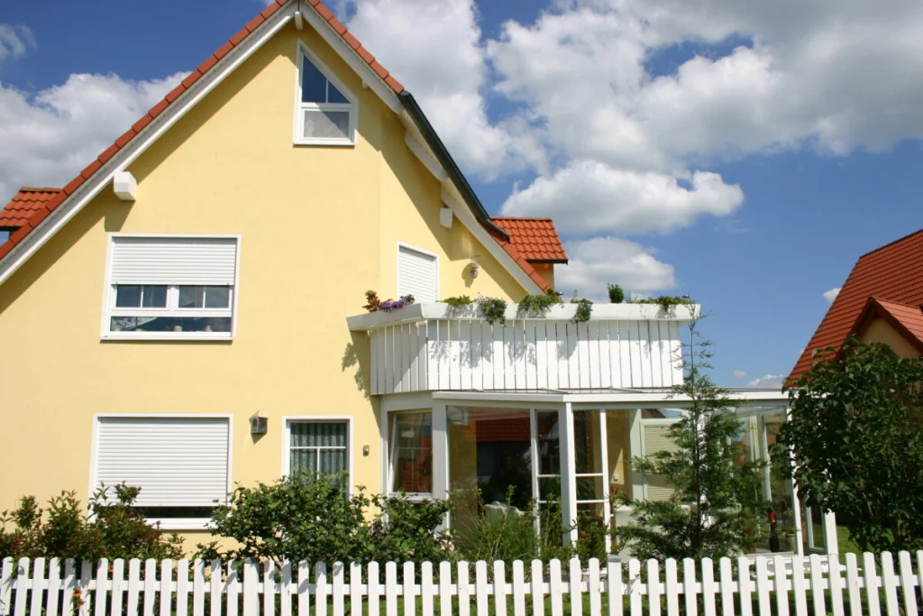 Gelbes Einfamilienhaus in Hausen mit weißem Wintergarten unter dem Eckbalkon, Blick über den Gartenzaun