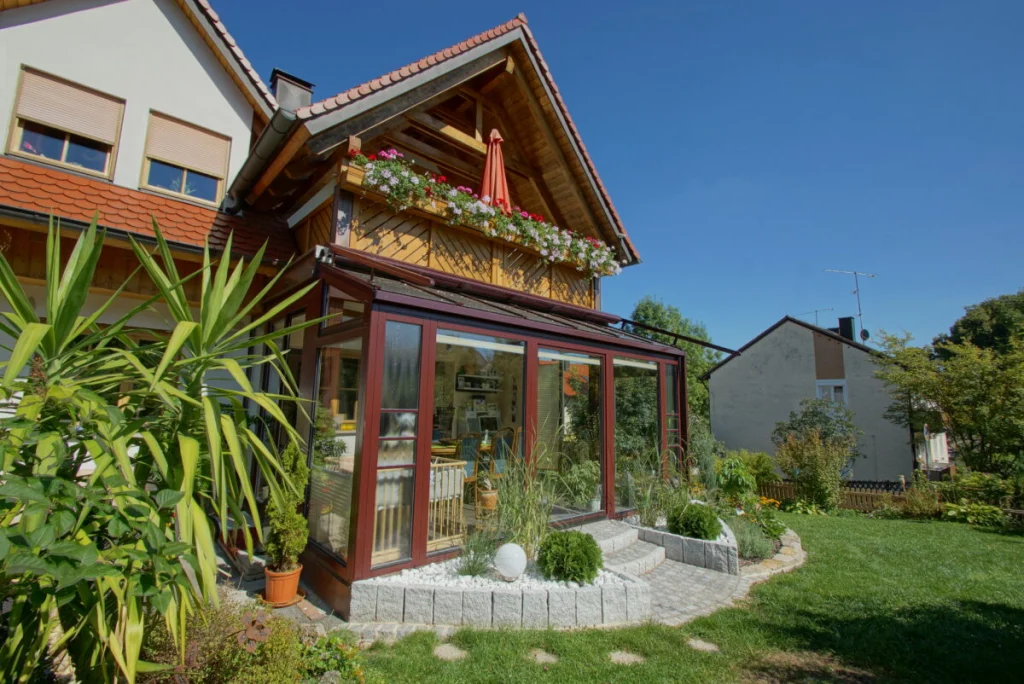 Wintergarten in Heiligenstadt mit dunkelroten Profilen unter dem Holzbalkon, Glasdach und gepflasterter Terrasse