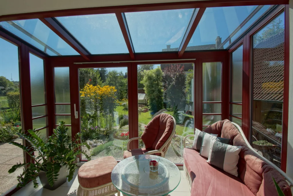 Wintergarten in Hormersdorf innen, Glasdach mit weinroten Sparren, Tür zum Garten, Korbsofa und Pflanzen