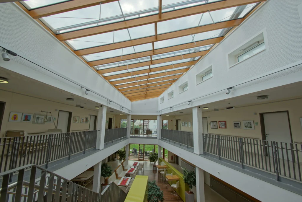 Atrium der Klinik Ebermannstadt mit Glasdach auf Holzsparren, Galerie im Obergeschoss und Sitzbereich unten