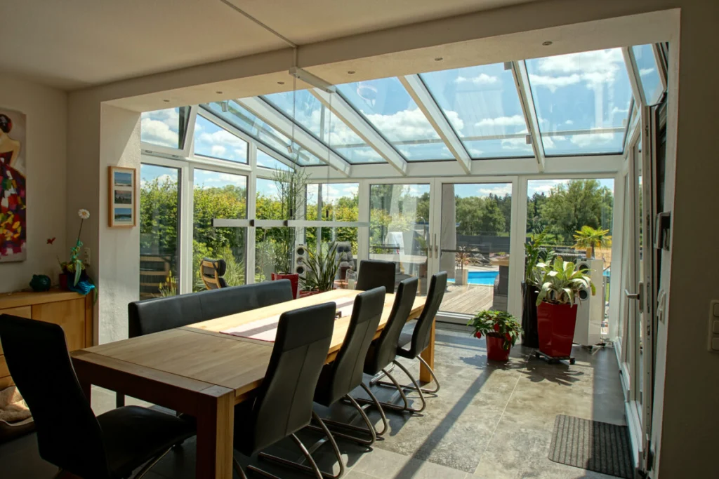 Wintergarten in Königsfeld mit Pultdach aus Glas, langem Esstisch und offener Tür zur Holzterrasse mit Pool