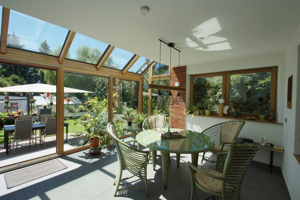 Wintergarten in Ahorntal innen, Holzkonstruktion mit Glasdach, grüne Rattanmöbel und offene Tür zur Terrasse