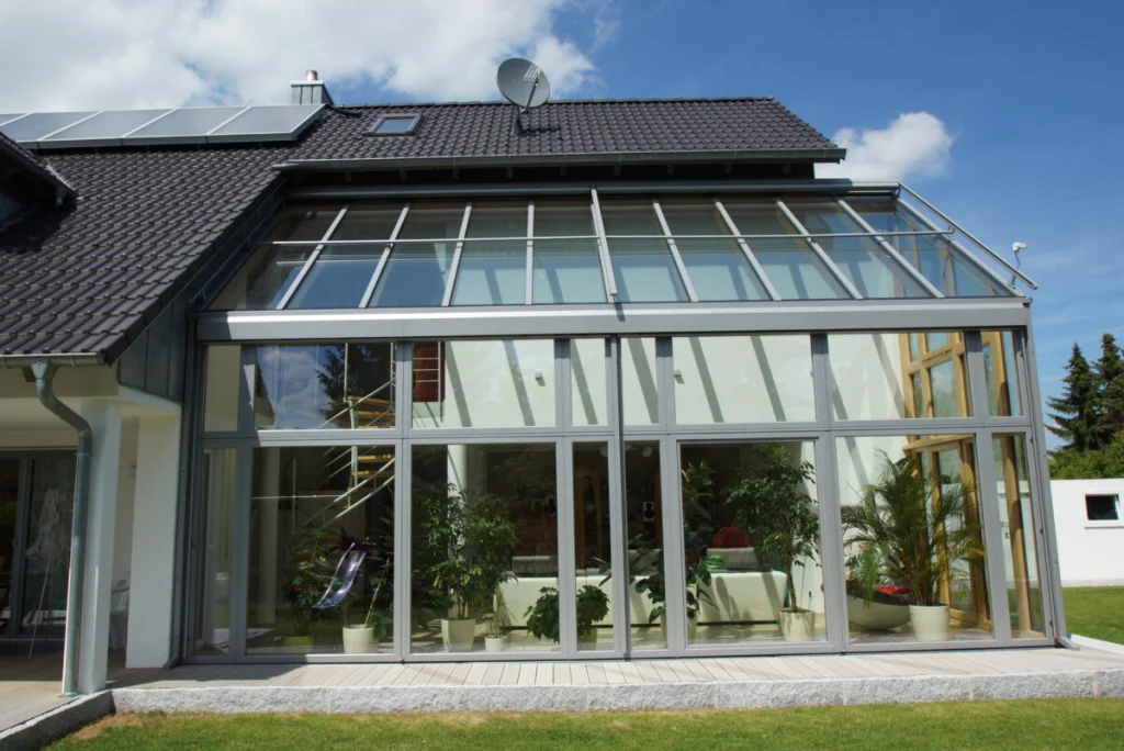 Glasanbau in Bruck von vorn, Glasdach und Glasfront mit hellgrauen Aluminiumrahmen, innen Wendeltreppe und Pflanzen