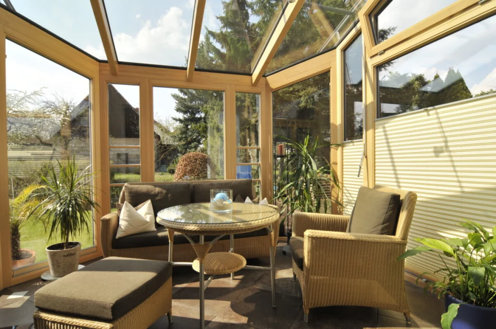 Wintergarten in Buch mit Rattanmöbeln, Meranti-Holzkonstruktion, Glasdach und Plisseestores, Blick auf Rasen und Bäume