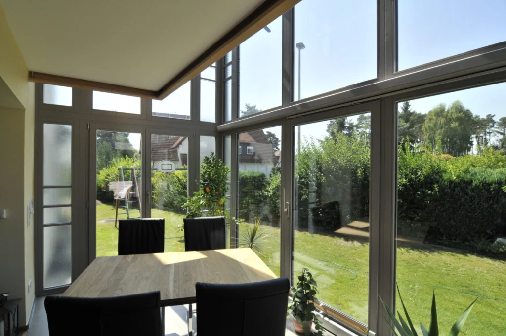 Wintergarten in Hannberg, Esstisch mit schwarzen Stühlen, Glasfassade mit grauen Profilen und Blick auf Rasen und Hecke