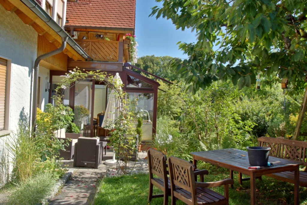 Wintergarten in Heiligenstadt von der Gartenseite mit dunkelroten Profilen, Rosenbogen und Gartenmöbeln
