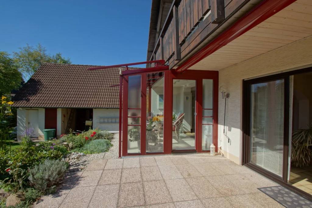 Wintergarten in Hormersdorf von der Terrasse, weinrote Rahmen unter dem Holzbalkon des Hauses, Glastür und Strukturglas