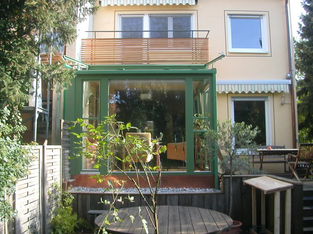 Wintergarten in Schoppershof mit grünen Rahmen unter dem Holzbalkon eines Reihenhauses, frontal vom Garten