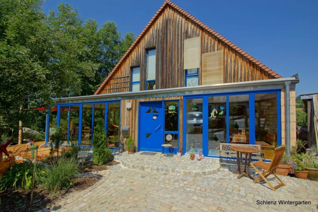 Flachdachanbau in Grafenau mit blauen Rahmen und blauer Haustür vor einem Holzhaus mit Satteldach, Pflasterhof davor