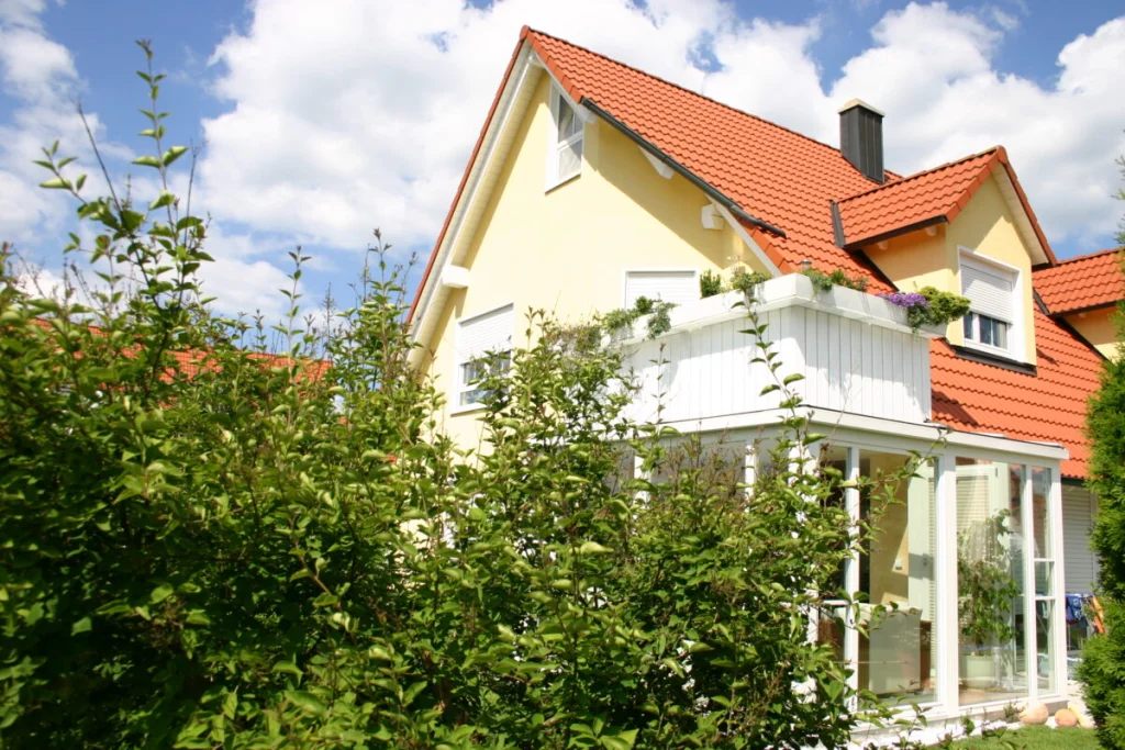 Wintergarten in Hausen unter dem Balkon, weiße Profile, gelbes Haus mit roten Ziegeln, Sträucher im Vordergrund