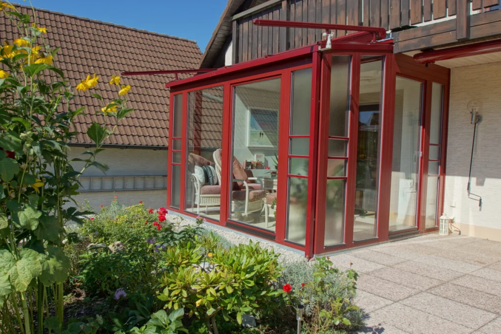 Wintergarten in Hormersdorf vom Garten, kleiner Anbau mit Pultdach und weinroten Rahmen, Korbmöbel innen, Blumenbeet davor