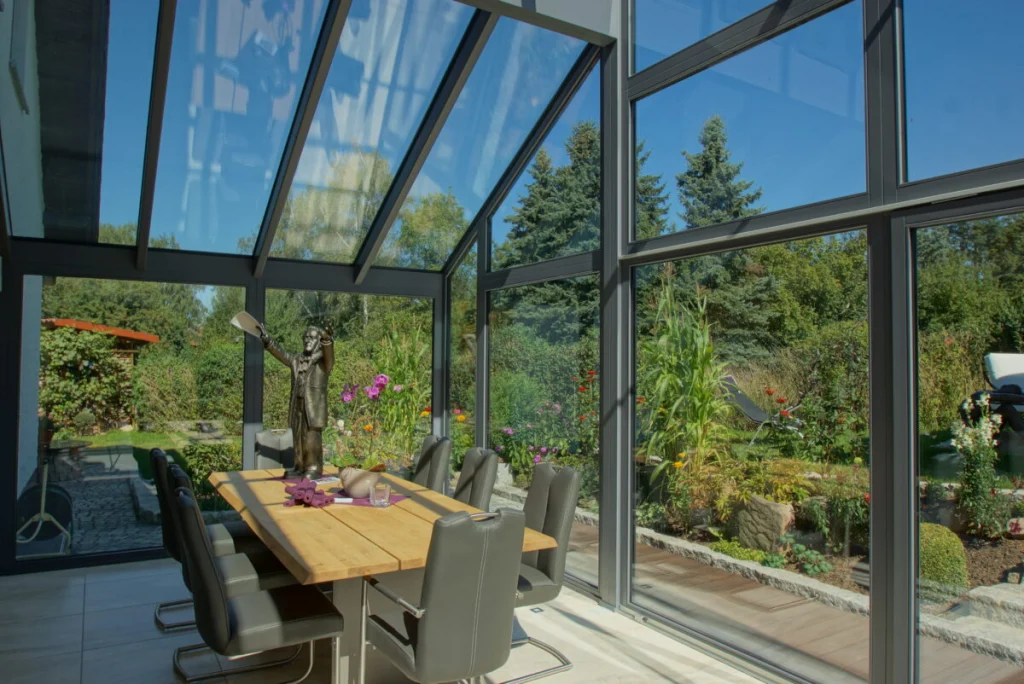 Wintergarten in Bayreuth, Glasgiebel mit grauen Profilen, Esstisch aus Massivholz und Blick in den Garten mit Fichten
