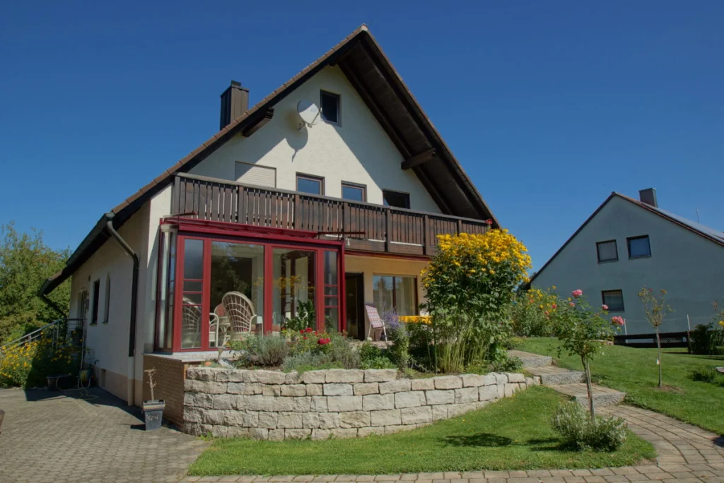 Haus in Hormersdorf mit Satteldach, dunklem Holzbalkon und weinrotem Wintergarten darunter, Natursteinmauer und Garten