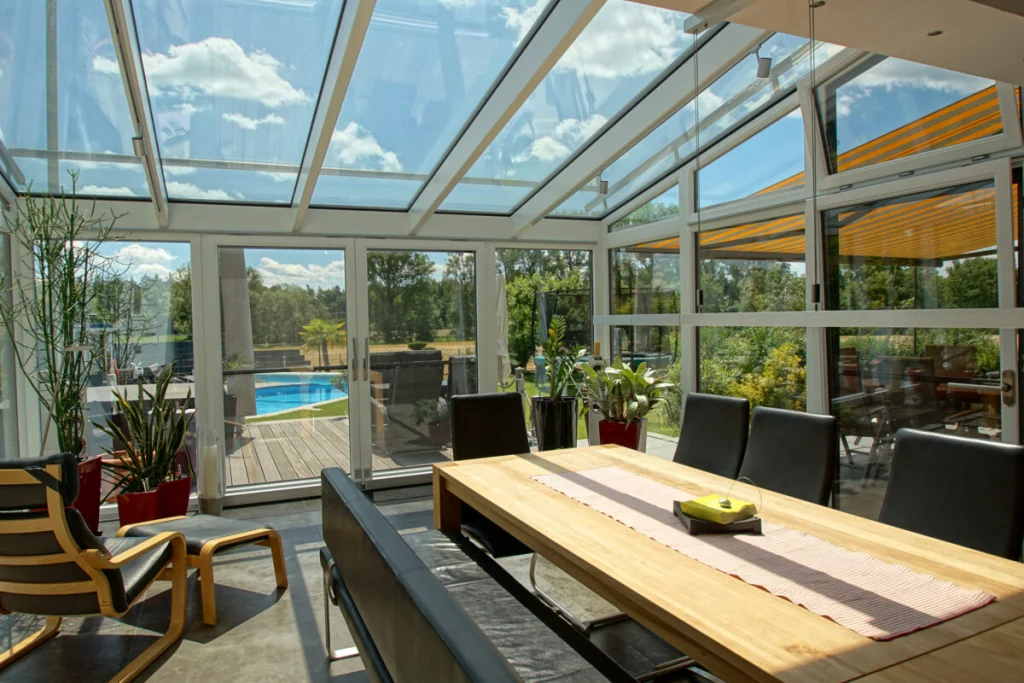 Wintergarten in Königsfeld mit Glasdach, Esstisch aus Massivholz und Blick auf Terrasse, Pool und Wiese