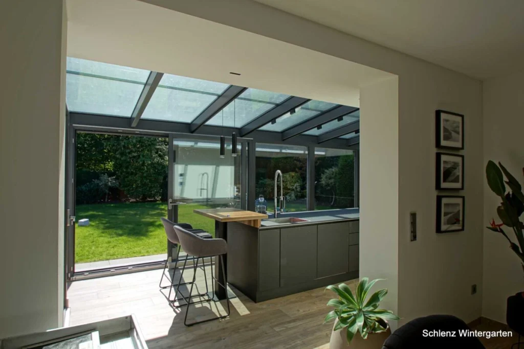 Wintergarten in Rathsberg innen, Küche mit grauer Kochinsel und Barhockern unter dem Glasdach, offene Tür zum Garten