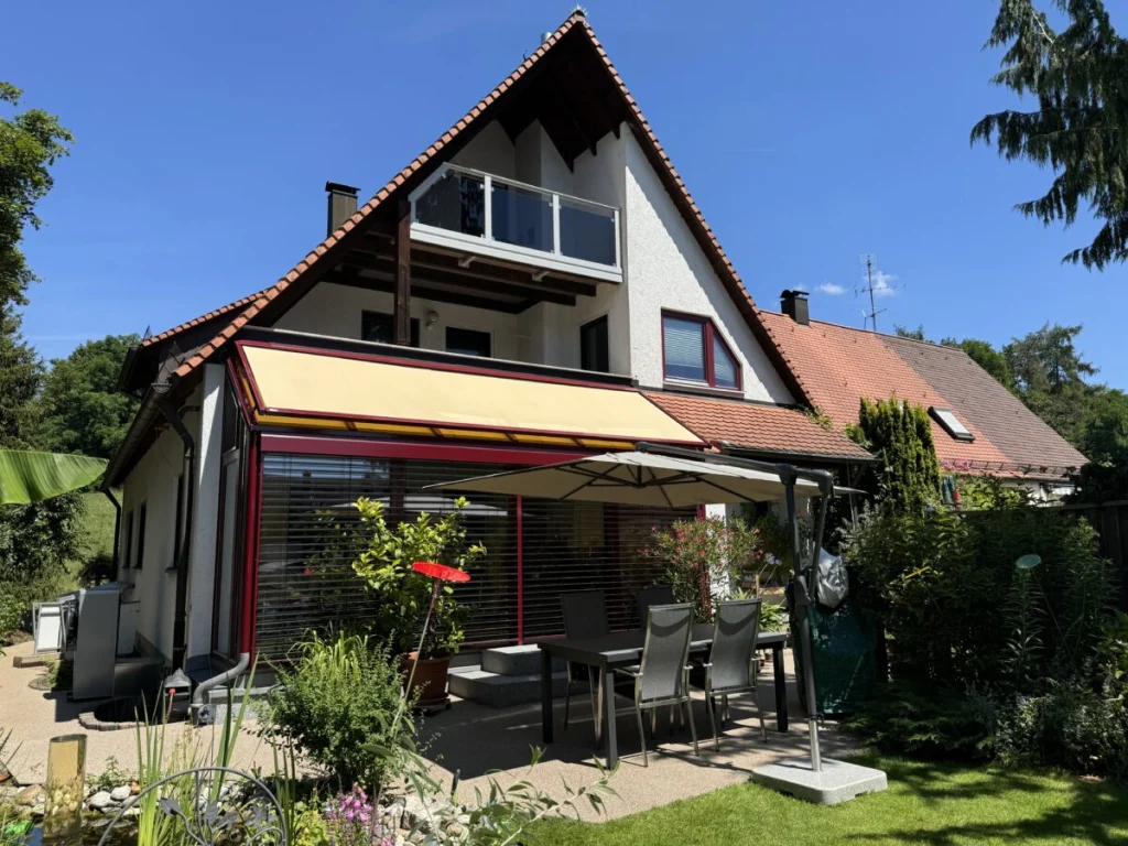 Wintergarten in Ahorntal unter dem Balkon, weinrote Profile, ausgefahrene Markise und Terrasse mit Esstisch