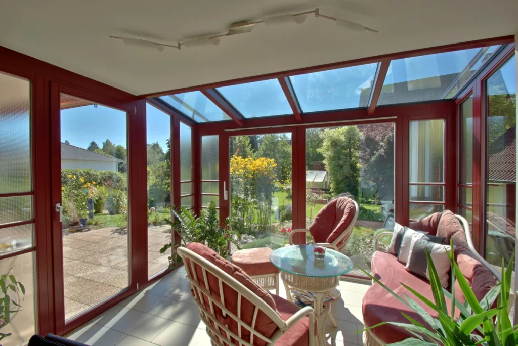 Wintergarten in Hormersdorf innen mit Glasdach, weinroten Rahmen, Korbsesseln und Tür zur Terrasse
