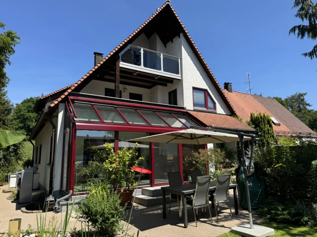 Wintergarten in Ahorntal von außen mit weinroten Profilen und Pultdach aus Glas unter dem Balkon, Terrasse mit Sonnenschirm
