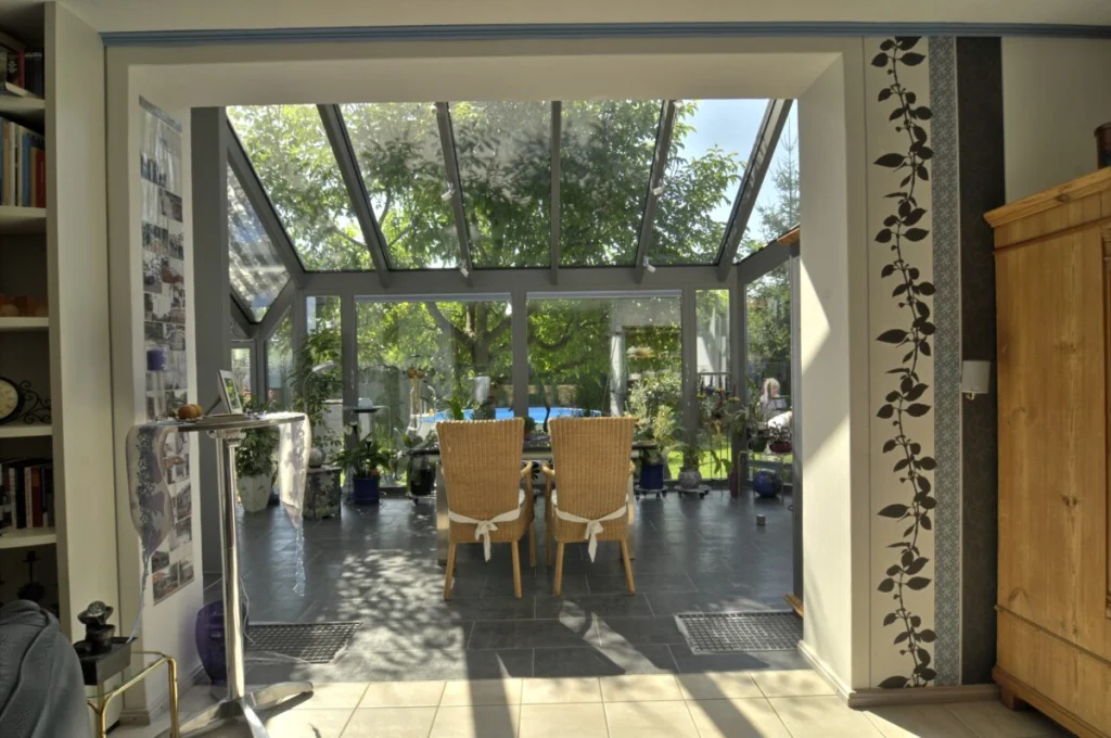 Blick aus dem Wohnraum in den Wintergarten in Kleinsendelbach, graue Profile, Glasdach, Rattanstühle und Garten