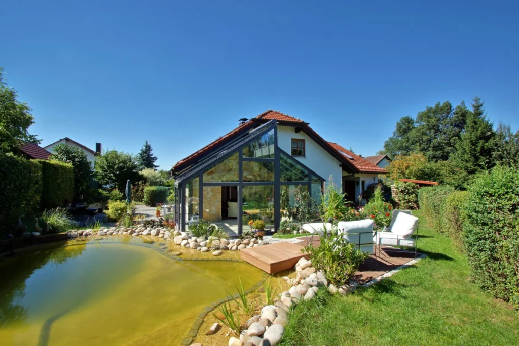Wintergarten in Bayreuth mit Satteldach aus Glas am Schwimmteich, Außenansicht mit Holzsteg und Gartenmöbeln