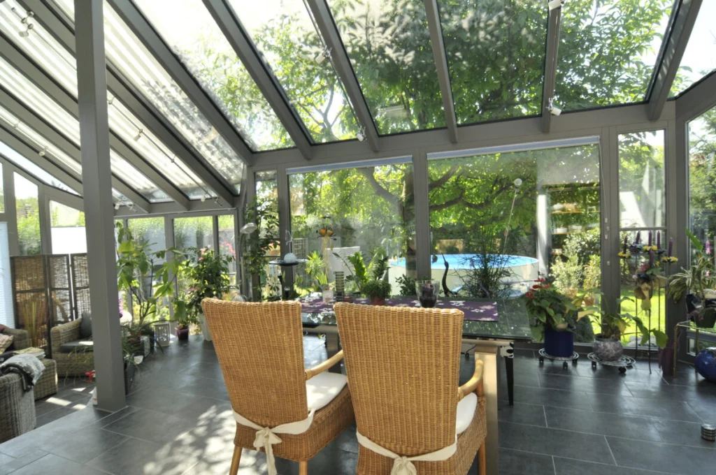 Wintergarten in Kleinsendelbach von innen mit Glasdach, zwei Rattanstühlen am Glastisch und Blick auf Baum und Pool