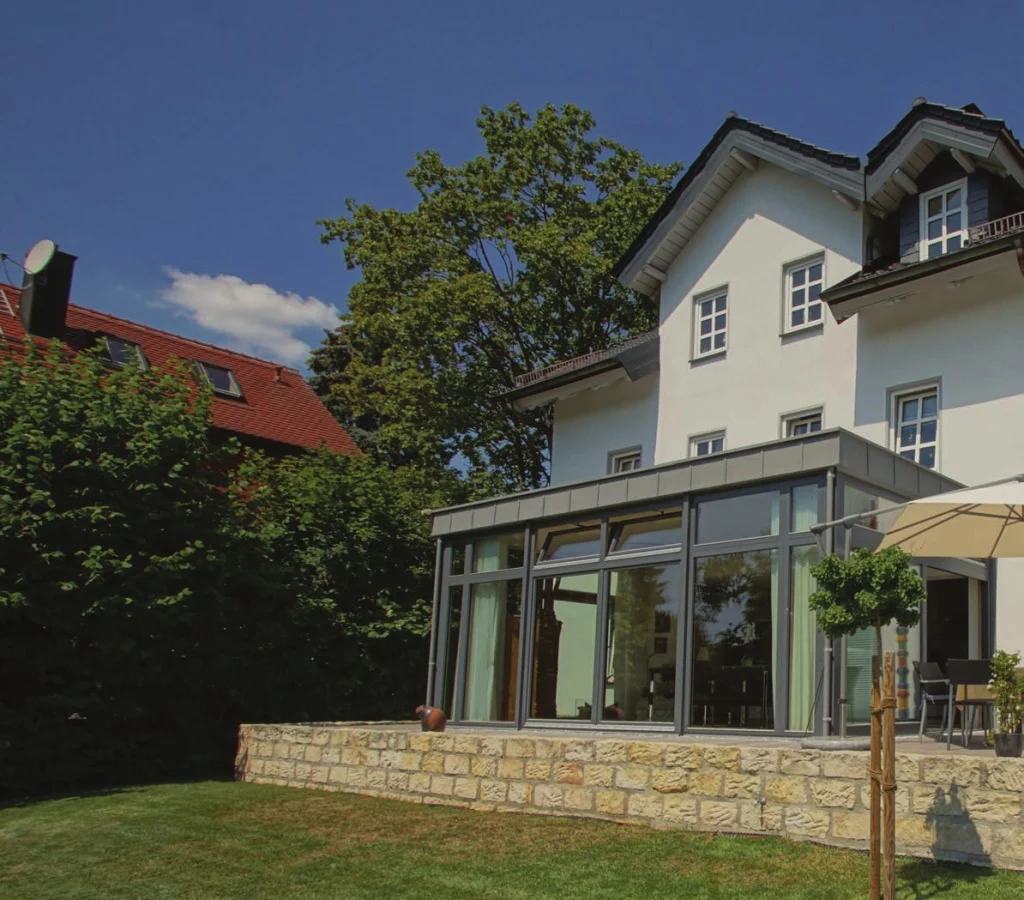 Klassische Villa mit verglastem Anbau als Esszimmer, Natursteinmauer und Sonnenschirm